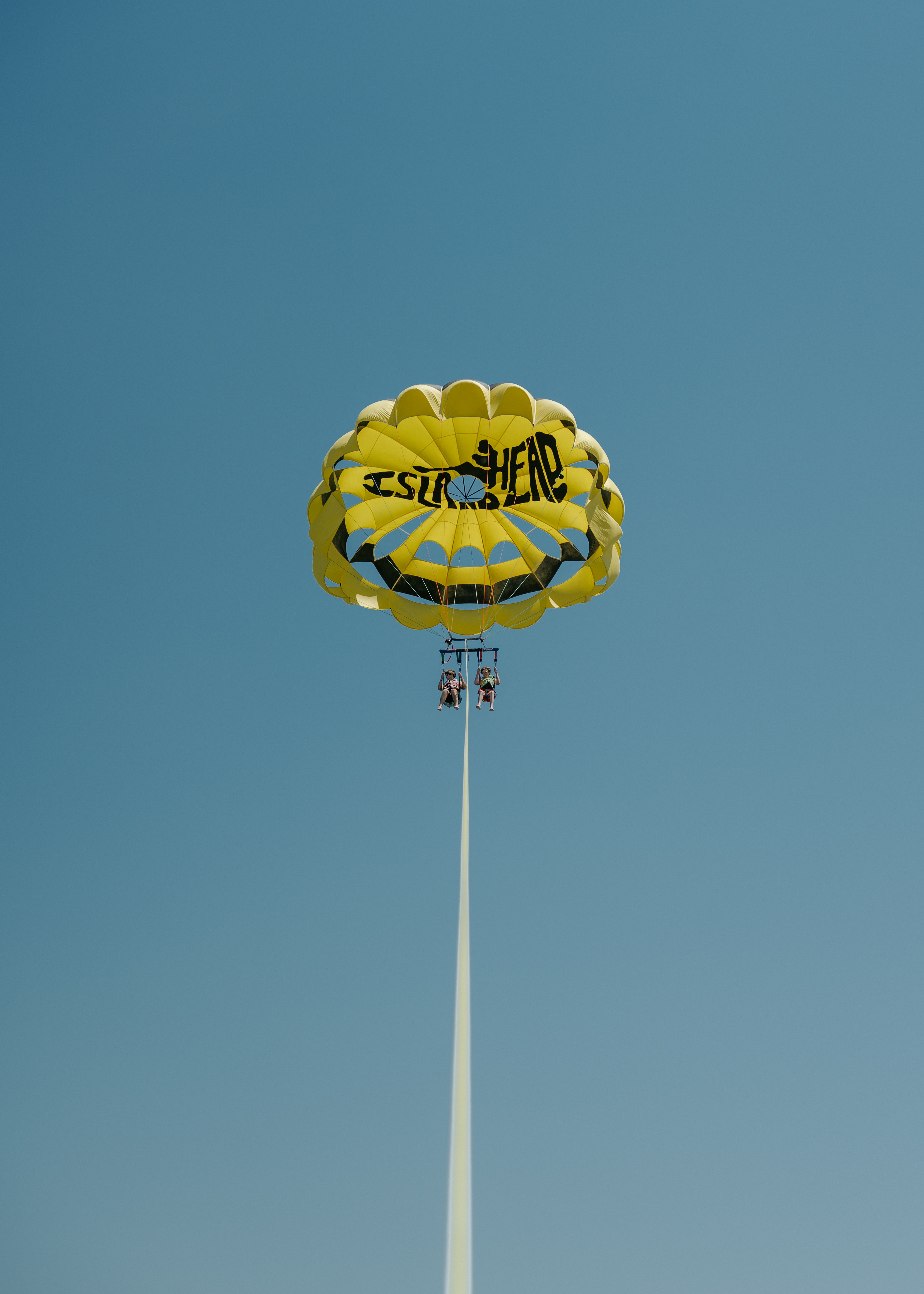 Bright yellow parasail with two riders suspended beneath it, towed on a long rope and soaring against a clear blue sky.