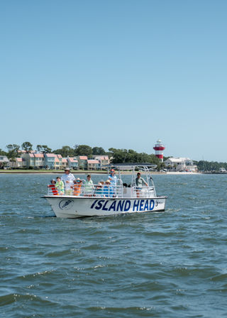 Sightseeing boat with passengers cruising a coastal bay past pastel beachfront cottages and a red-and-white lighthouse on a sunny day.