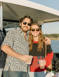 Smiling friends in sunglasses enjoying drinks on a sunny boat ride along a calm shoreline with blue sky and casual summer outfits.