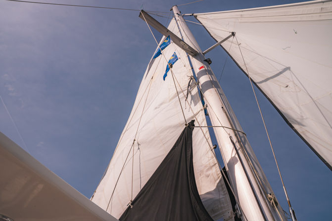 Looking up a sailboat mast with rigging, billowing white sails and a black sail cover, small blue signal flags flying against a clear blue sky