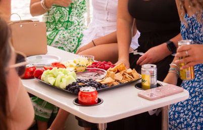 Summer boat party snack tray with honeydew, blueberries, strawberries, raspberries, pineapple, chips and canned drinks on a white table surrounded by friends in sundresses.