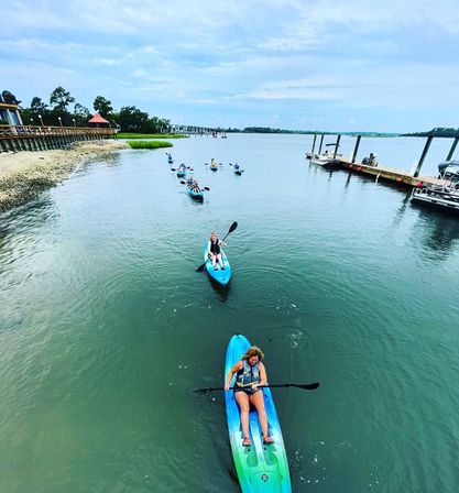 Group of kayakers paddling blue and green sit-on-top kayaks along a calm coastal inlet past a shoreline boardwalk and marina docks under a cloudy sky