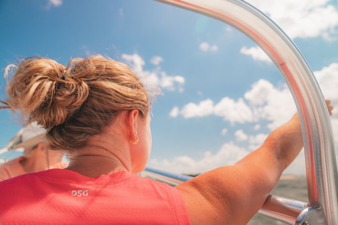Back view of a sun-soaked woman with a messy bun and gold hoop earring in a coral shirt holding a boat railing over open water under a bright blue sky with fluffy clouds.