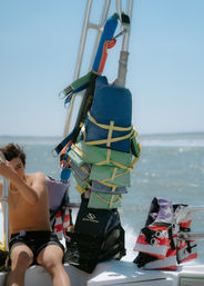Summer boat scene with colorful life jackets and safety vests stacked on the frame, an American-flag swim vest on the seat, a passenger holding the rail and sunlit ocean on the horizon.