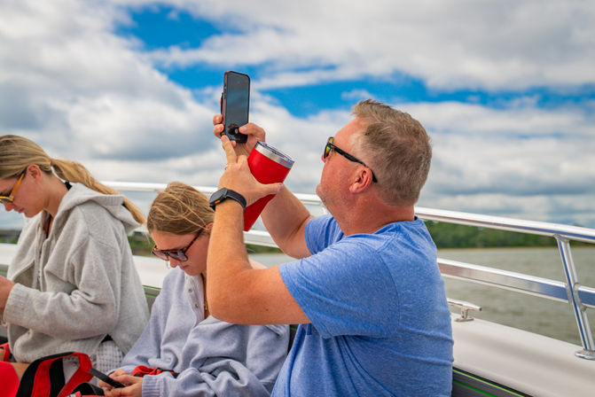 Family on a boat on a lake: man in blue shirt and sunglasses lifts a smartphone and red travel tumbler to take a photo while two passengers scroll phones under a cloudy sky and treelined shore.