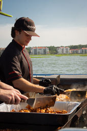 Cook in black uniform using spatulas to grill shrimp and seasoned chicken on a large outdoor flat-top by a scenic waterfront with coastal condo buildings in the background.
