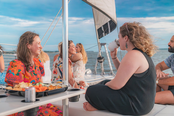 Group of friends socializing on a sailboat cruise — colorful dress, snack platter and drinks on the table, one person sipping from a tumbler as the boat sails on calm ocean under a blue sky.