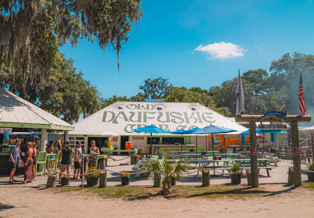 Sunny coastal outdoor seafood bar with a white barn-style roof painted sign, picnic tables and blue umbrellas, diners in line, and live oak trees draped with Spanish moss for casual beachfront dining.