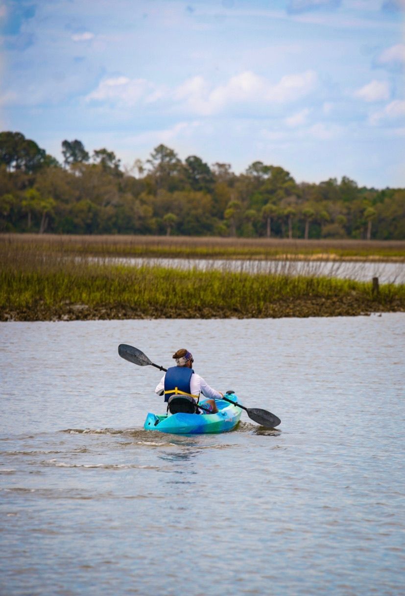 Solo paddler in a turquoise kayak gliding through a coastal marsh estuary with tidal grasses and a tree-lined shoreline under a partly cloudy sky