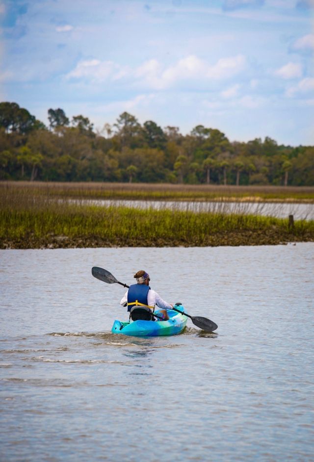 Solo paddler in a turquoise kayak gliding through a coastal marsh estuary with tidal grasses and a tree-lined shoreline under a partly cloudy sky