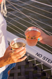 Close-up of two hands clinking plastic cups of sparkling rosé and champagne on a sailboat deck over water, with rope railings and a visible “NO STEP” sign — casual boating cheers.