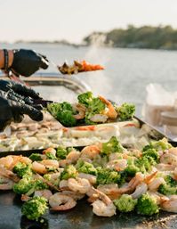Sunlit waterfront grill: sizzling shrimp and broccoli on a flat-top griddle, gloved hands serving fresh seafood at an outdoor cookout