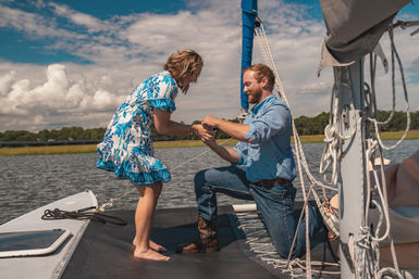 Romantic sailboat proposal: man kneeling to present a ring to a woman in a blue floral dress on calm coastal waters with marshland and a partly cloudy sky.