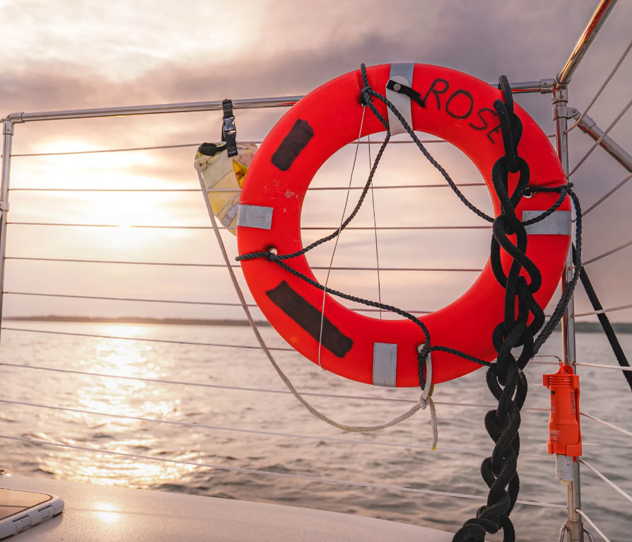 Bright red lifebuoy labeled 'ROSE' with coiled black rope attached to a sailboat railing, set against a golden sunset reflecting on calm open water.