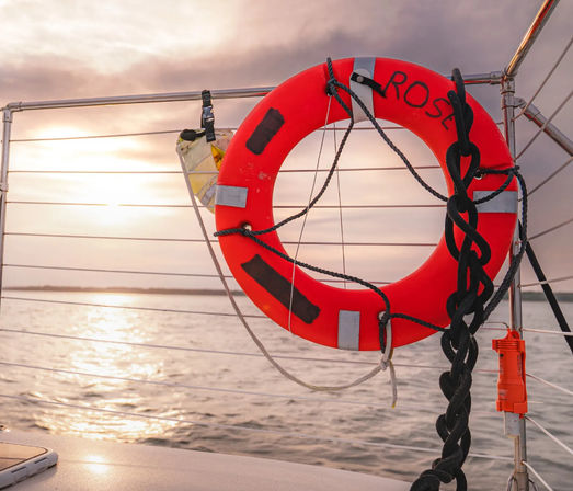 Bright red lifebuoy labeled 'ROSE' with coiled black rope attached to a sailboat railing, set against a golden sunset reflecting on calm open water.
