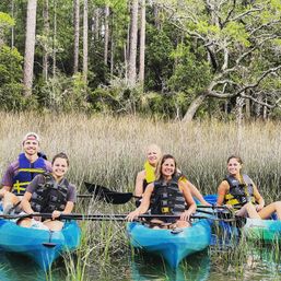 Five smiling kayakers in blue kayaks wearing life jackets paddling through a coastal salt marsh with tall grasses and pine trees