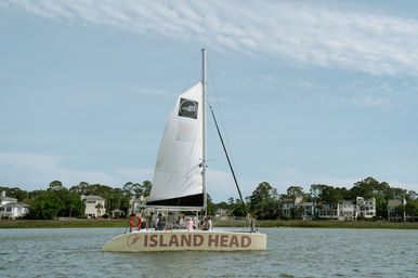 Leisure sailboat gliding across a calm coastal inlet with passengers onboard, passing waterfront homes and marsh under a bright blue sky.