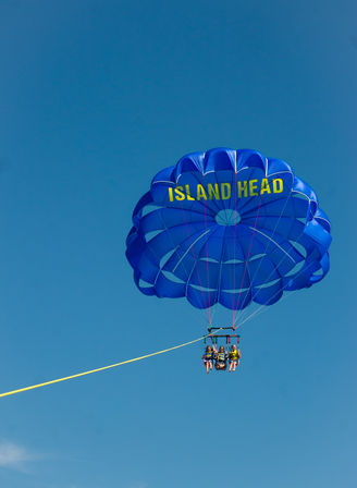 Four thrill-seekers parasailing under a large blue canopy with yellow lettering, suspended by a yellow tow rope against a clear blue sky.