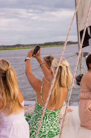 Three friends on a sailboat along a coastal waterway, one holding a smartphone up to photograph the shoreline; nautical ropes, sail, and casual summer dresses visible.
