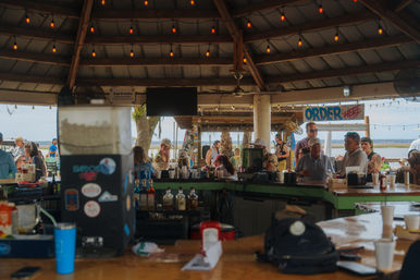 Busy waterfront tiki-style outdoor bar with patrons lined along a wooden counter under a string-lit pavilion, 'Order Here' sign and marshy water view in the background