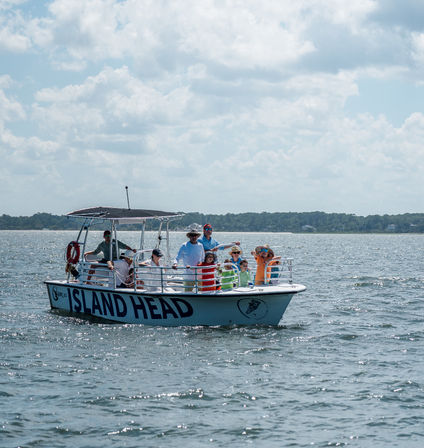 Sightseeing boat tour with adults and kids on a sunny summer day in a calm coastal bay, passengers wearing colorful life jackets and sun hats under a partly cloudy sky.