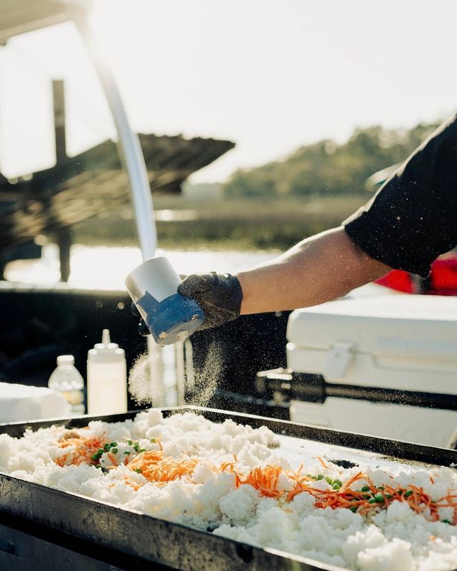 Gloved hand sprinkling seasoning over a sunlit outdoor griddle of rice with shredded carrots and peas at a waterfront food stall.