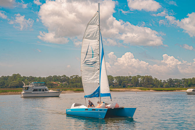 Bright blue catamaran with white sails gliding across calm coastal waters for a leisurely sail on a sunny, partly cloudy day, passengers relaxing on deck with tree-lined shoreline and other boats in the background.
