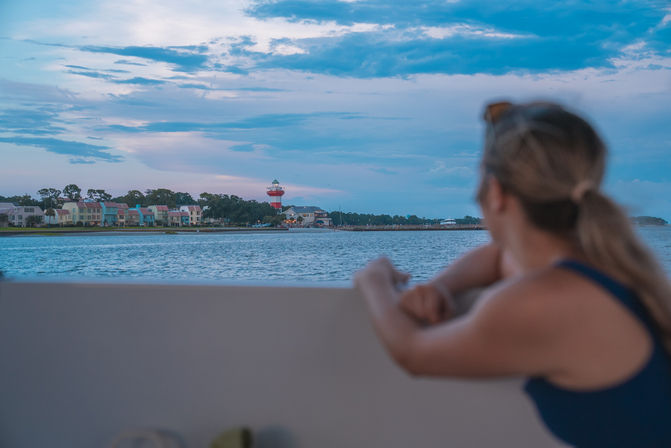 Person leaning on a boat railing, gazing across a calm bay toward a coastal town with pastel waterfront houses and a red-and-white striped lighthouse under a pastel blue dusk sky.