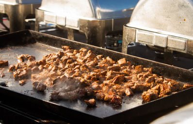 Juicy grilled beef cubes sizzling on a flat-top griddle with steam rising, framed by chafing trays — outdoor catering or street-food station.