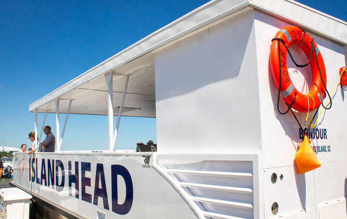 White island ferry docked at a sunny coastal harbor with passengers on the open upper deck, large blue lettering on the hull and a bright orange lifebuoy against a clear blue sky.
