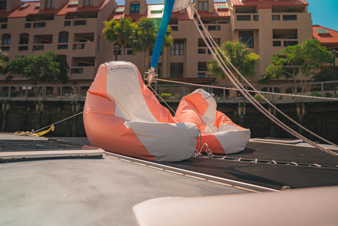Two orange-and-white inflatable lounge chairs on a sailboat deck with rigging ropes, palm trees and waterfront apartment buildings at a sunny marina
