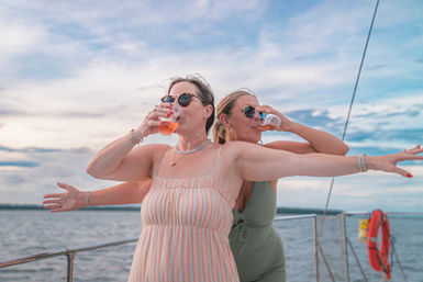 Two friends on a sailboat wearing sunglasses and summer outfits, arms out like Titanic while sipping drinks with the ocean and cloudy sky in the background.