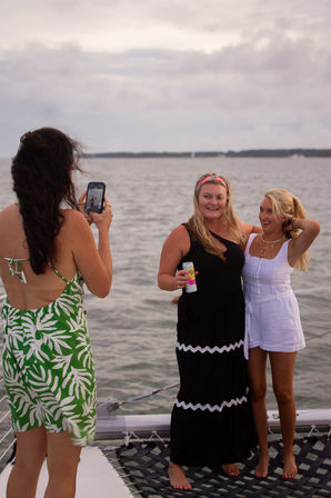 Three friends posing barefoot on a sailboat trampoline over calm coastal water with a cloudy sky — one woman in a green tropical dress snaps a photo while a woman in a black dress holds a canned drink and a woman in a white romper smiles.