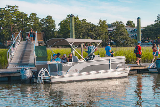 Pontoon boat with canopy docked at a wooden pier in a coastal marsh, passengers chatting and boarding on a sunny day with grassy shoreline and nearby pedestrians.
