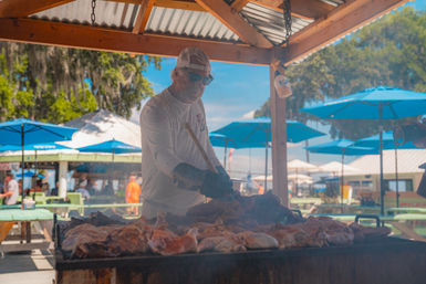 Smoky outdoor barbecue with a cap-wearing cook tending a large pit of roasting meats under a wooden pavilion, blue patio umbrellas and picnic tables on a sunny day