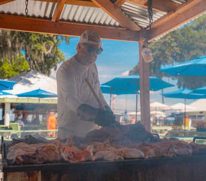 Smoky outdoor barbecue with a cap-wearing cook tending a large pit of roasting meats under a wooden pavilion, blue patio umbrellas and picnic tables on a sunny day