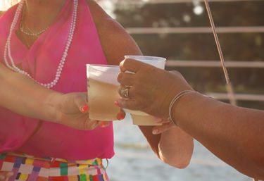 Two people clinking frosty plastic cups at a beachside sunset, hands with rings and bracelets, bright pink top and colorful patterned skirt visible