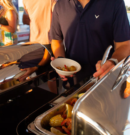 Person serving a shrimp boil with corn, potatoes and lemon from a chafing dish at an outdoor waterfront buffet on a boat