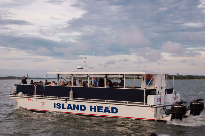Passenger sightseeing ferry with open deck and passengers cruising coastal waters near a tree-lined shore under a dramatic cloudy sky