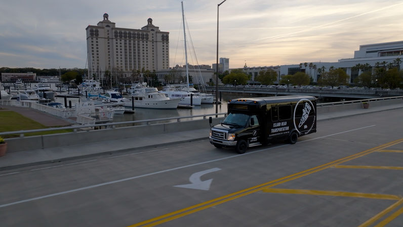 Black shuttle bus driving across a waterfront bridge past a marina of yachts and a tall hotel at sunset