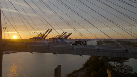 Golden sunset over a cable-stayed bridge spanning a river, silhouetted container cranes at a busy port and a truck crossing with shimmering water reflections