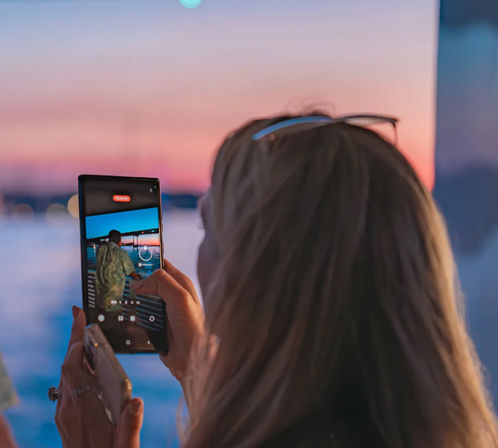 Person seen from behind holding a smartphone to capture a colorful waterfront sunset, phone screen showing the camera app framing a silhouetted figure against a pastel sky and calm water.