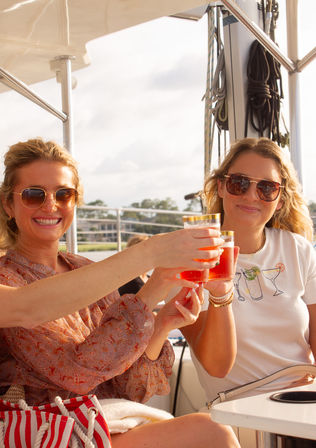 Two friends clinking bright red cocktails on a sunny sailboat deck near a coastal marina, wearing sunglasses and casual summer outfits