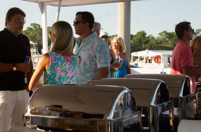 Group of adults in colorful summer outfits socializing around buffet chafing dishes on a sunny boat deck near a coastal harbor.