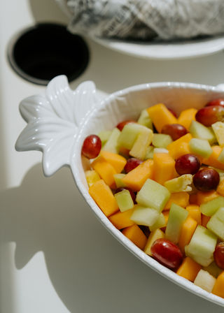 Close-up of sunlit fresh fruit salad with diced cantaloupe and honeydew melon and red grapes in a white leaf-shaped ceramic bowl on a table
