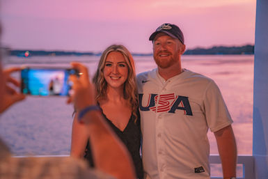 Smiling man in a USA baseball jersey and woman posing for a phone photo on a waterfront deck at a pink sunset over the bay.