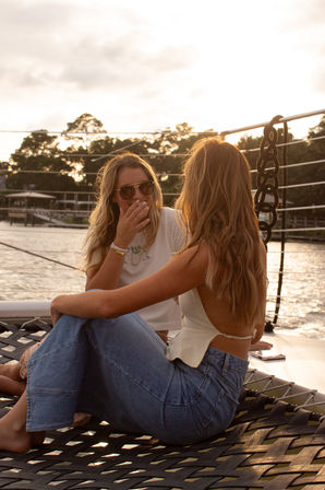 Two friends chatting on a sailboat's netted deck at sunset, relaxed summer vibe with waterfront docks and trees in the background.