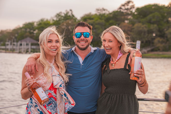 Three friends laughing on a boat deck during a sunset waterfront cruise, two women holding bottles of rosé and a man in sunglasses with his arms around them, marshy shoreline and docks in the background
