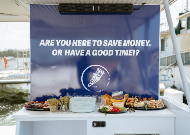 Appetizer buffet of plates, sandwiches, veggies and charcuterie on a boat counter, set against a large blue sign asking 'Are you here to save money, or have a good time!?', with marina boats and water in the background.