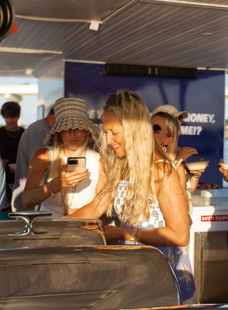 Sunlit summer yacht scene: smiling woman with long blonde hair serving from a buffet chafing dish on a boat deck while friends in sun hats and sunglasses check a phone during a coastal sunset party.